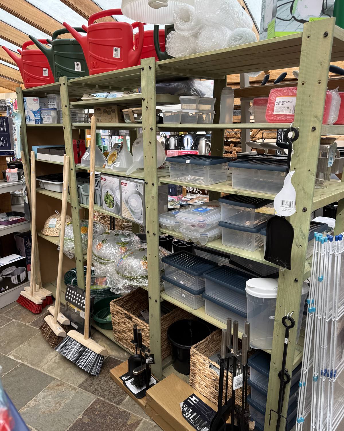 Green shelving bay of hardware: red watering cans, storage boxes, buckets and brooms